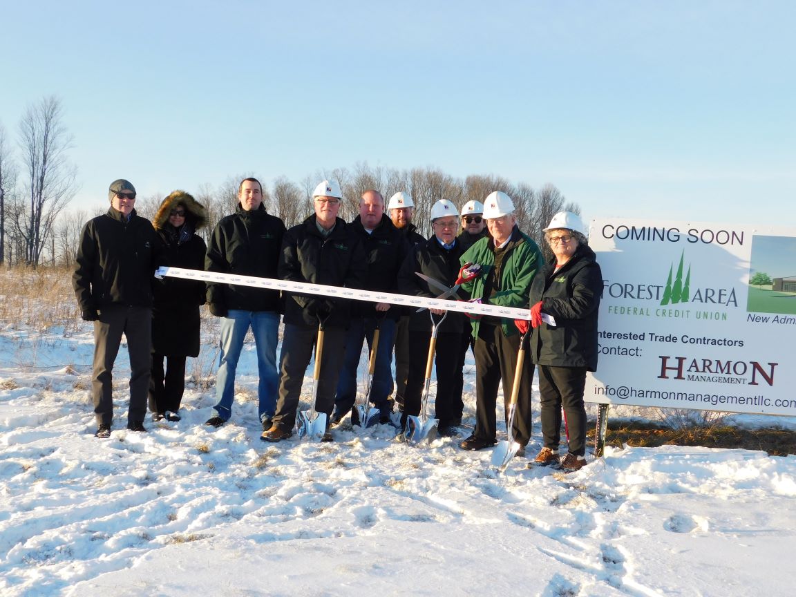 a group on Forest Area Federal Credit Union employees pose for a picture at the ground breaking for a new branch location. All are wearing hard hats and the sign reads "Coming Soon".