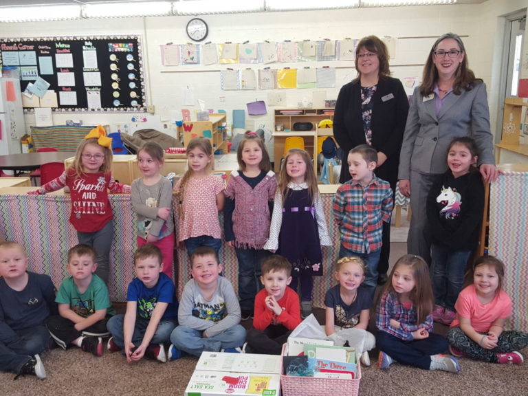 Two employees of Forest Area Federal Credit Union pose with a group of school aged children in their classroom. Everyone is smiling. 