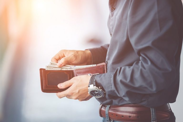 The torso of a man in a dress shirt and wearing a nice watch. He holds his wallet open and is pulling two one-hundred dollar bills from it. 