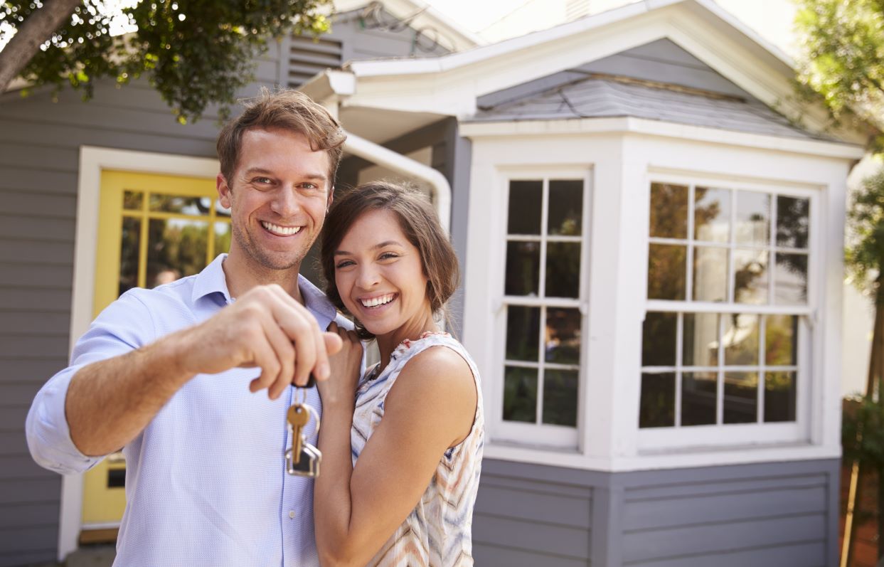 a young couple smile and holds keys to their new house while standing in front of the home. 