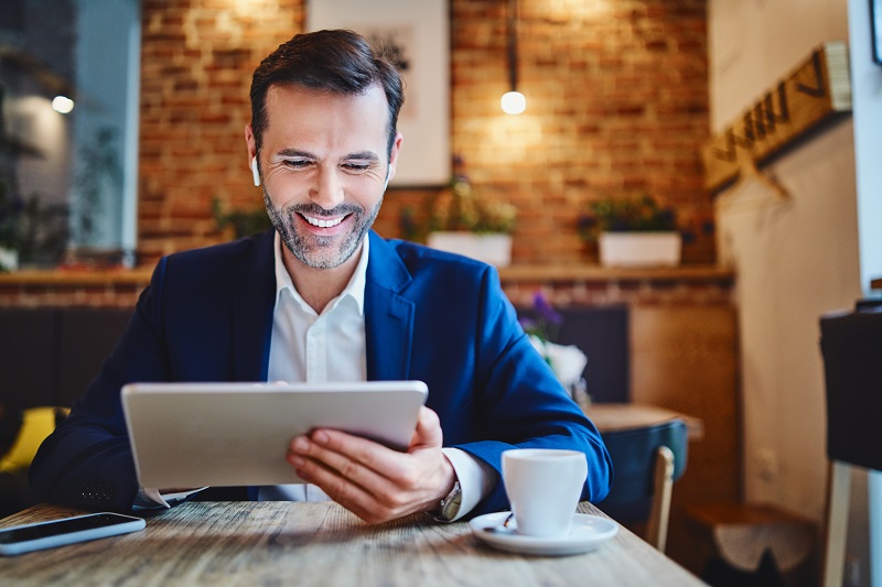 a middle aged man sitting at a coffee shop uses his tablet to live video chat with a Forest Area Federal Credit Union employee via Evergreen.